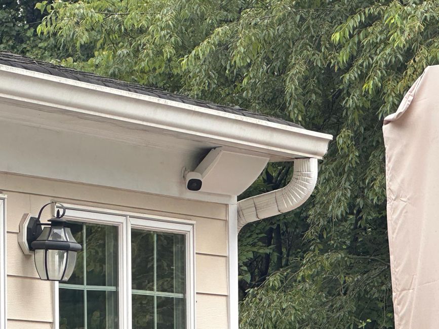Close-up of a white smart security camera mounted discreetly under the roof eave of a light yellow residential home.