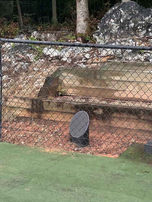 A dark grey cylindrical outdoor speaker positioned in the dirt behind a black chain-link fence, next to a wooden retaining wall.