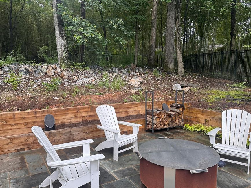 Outdoor patio area featuring white Adirondack chairs, a metal fire pit, a firewood rack, and landscape speakers against a wooded backdrop.