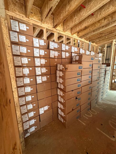 Large stack of brown cardboard shipping boxes with white labels stored at a construction site