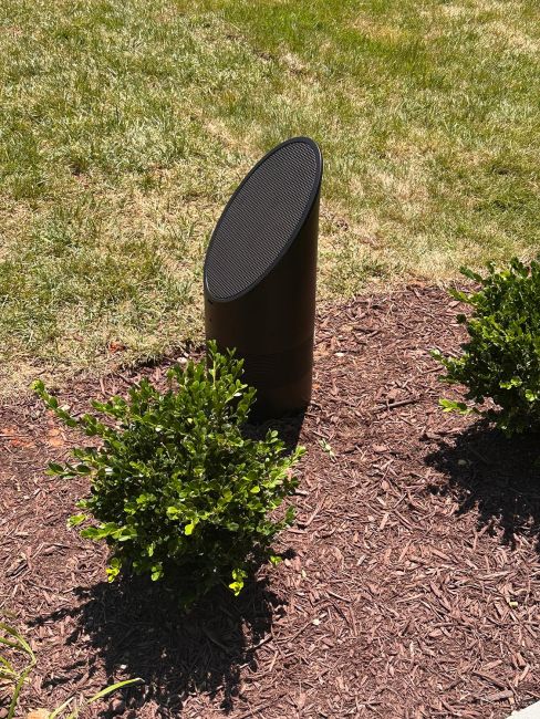 Close-up of a premium black cylindrical outdoor landscape speaker nestled in a mulch bed between green bushes on a sunny lawn.