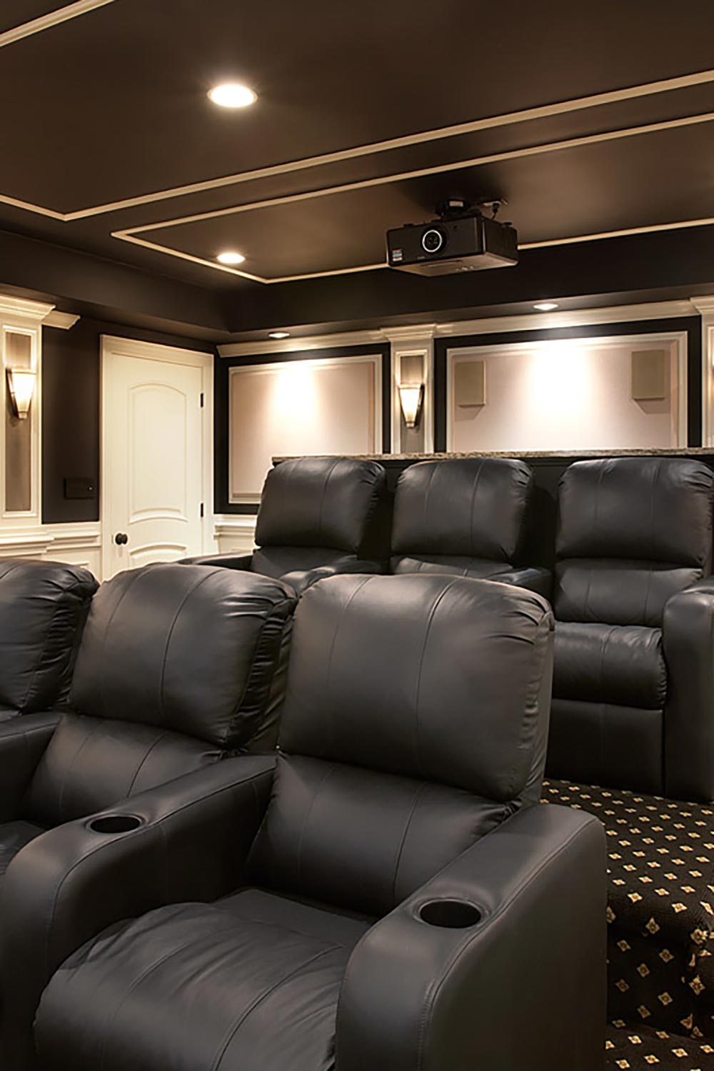 Black home theater projector mounted on a dark ceiling above rows of black leather seats