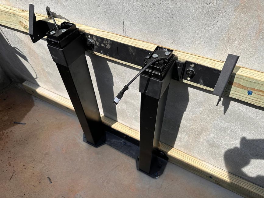 Close-up view of the black dual-column motorized mechanical tracks for an outdoor hidden TV lift system mounted to a wood frame inside a concrete vault.