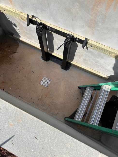 High-angle view inside the empty concrete underground TV lift bunker showing the black heavy-duty mounting tracks, a floor drain, and a green ladder.