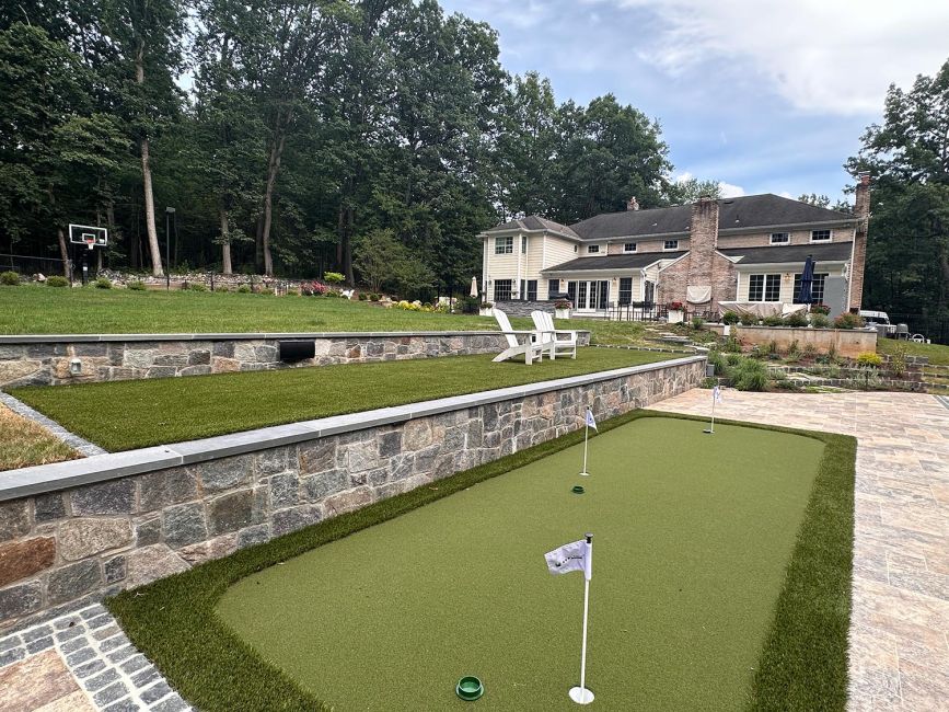 Wide shot of a multi-tiered luxury backyard featuring an artificial turf putting green, stone retaining walls, a swimming pool, and a large two-story house.